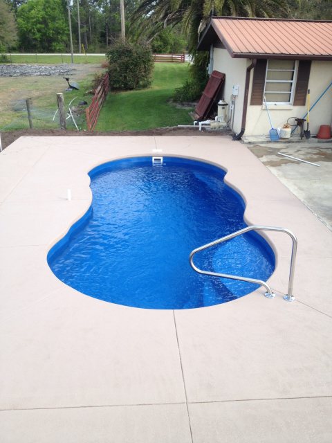 Fiberglass freeform pool with deep blue water and beige deck in a sunny backyard